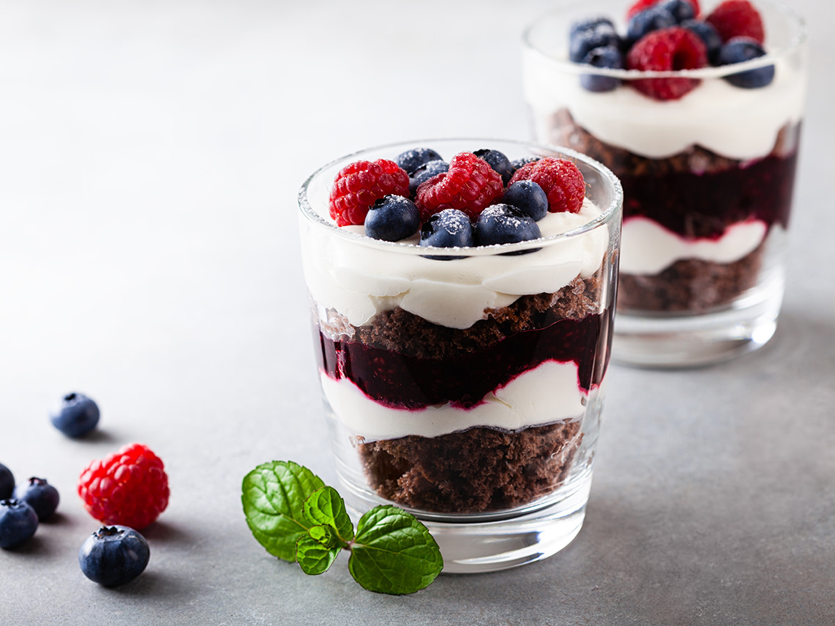 Auf einem hellen Hintergrund stehen zwei Desserts im Glas. Links daneben wurden Heidelbeeren und eine Himbeere drapiert. Henkelmann Gastronomiebetrieb