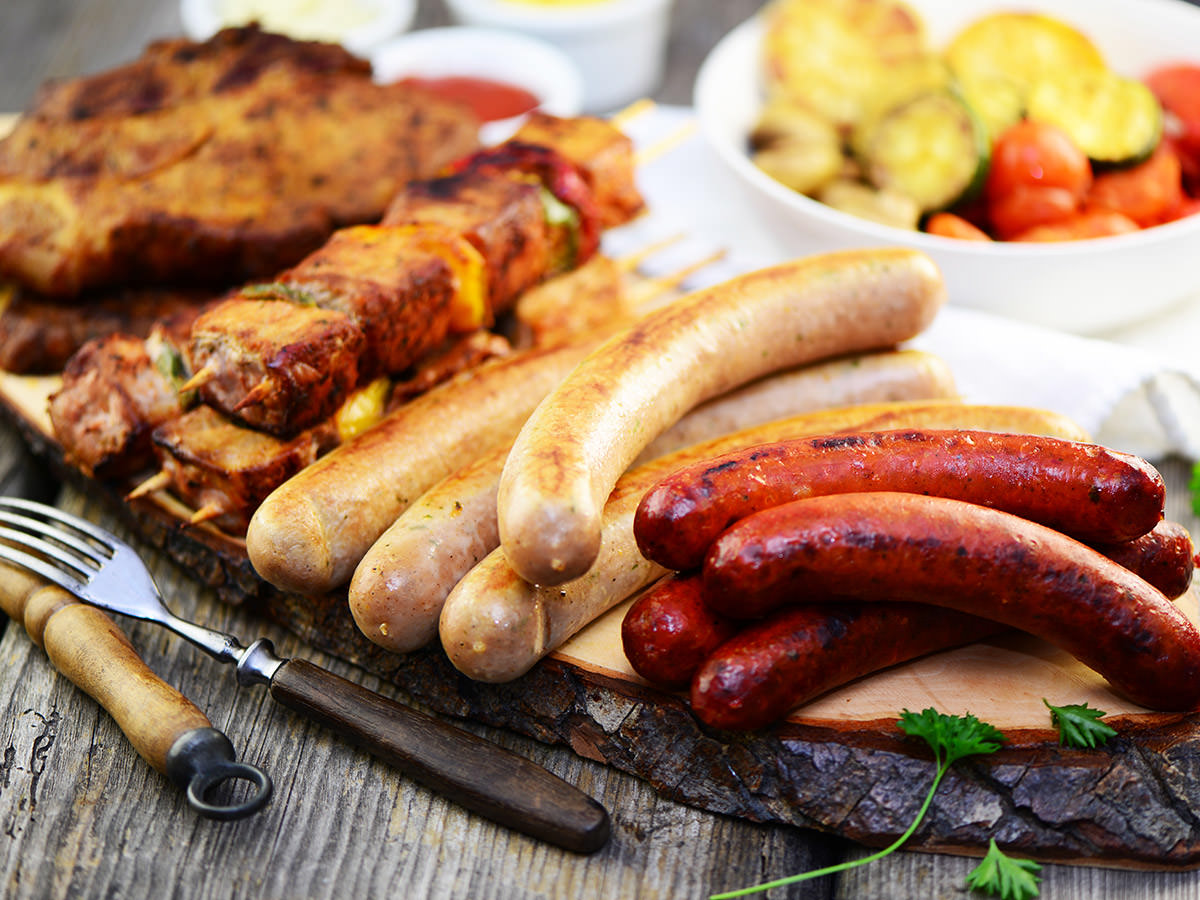 Auf einem rustikalen Tablett liegen Würstchen und Spieße. Links daneben befindet sich eine Gabel. Henkelmann Gastronomiebetrieb