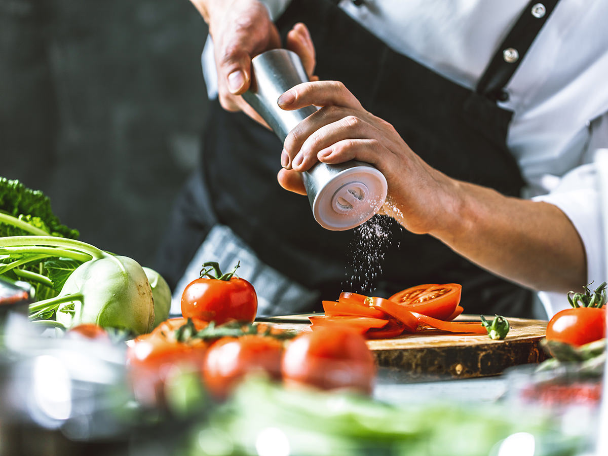 Ein Koch würzt die geschnittenen Tomaten mit einem Salzstreuer, die auf einem Schneidebrett liegen. Henkelmann Gastronomiebetrieb